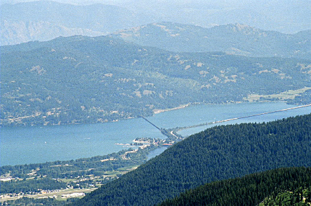 [Looking down the hill to the sliver of lake visible in this location. There are lots of evergreen covered low mountains on the other side of the bridge. The bridge curves as it goes across the water. A railroad section goes straight across a short section while the rest of the bridge turns to connect another shore.]